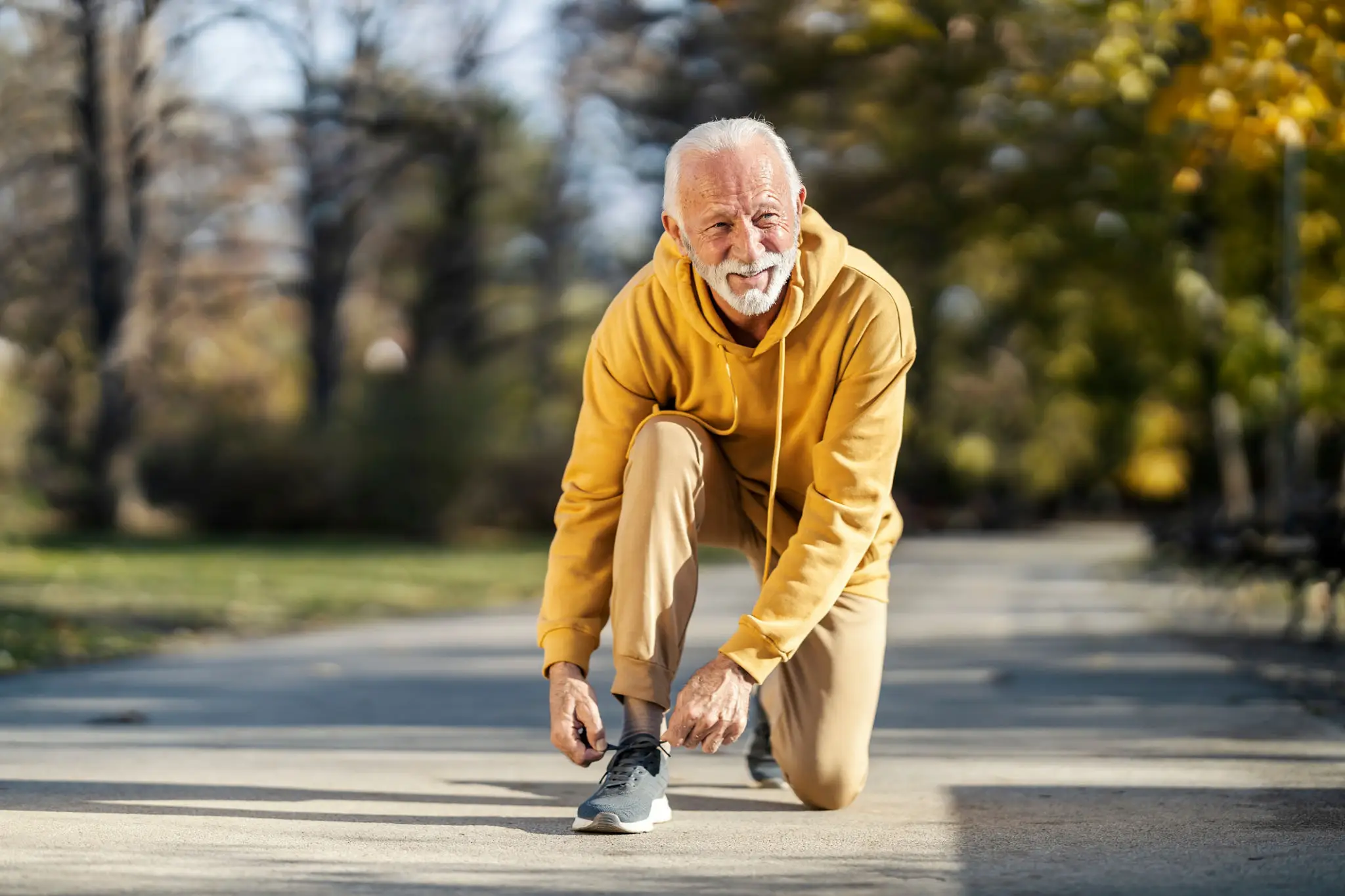 Elderly man tying his shoelaces outdoors, dressed in a yellow hoodie.