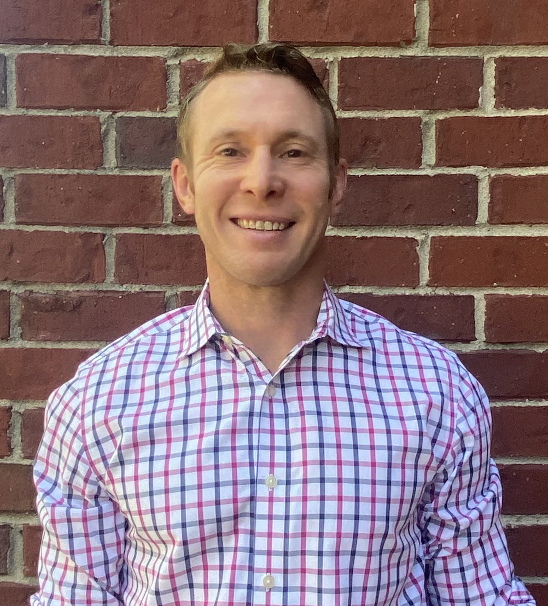 Smiling man in a checkered shirt standing against a brick wall.