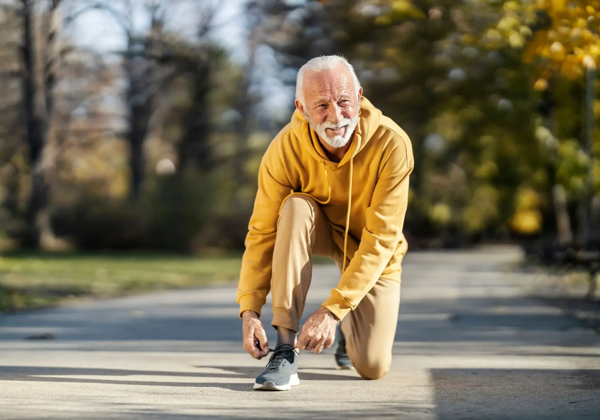 Elderly man tying his shoelaces outdoors, dressed in a yellow hoodie.
